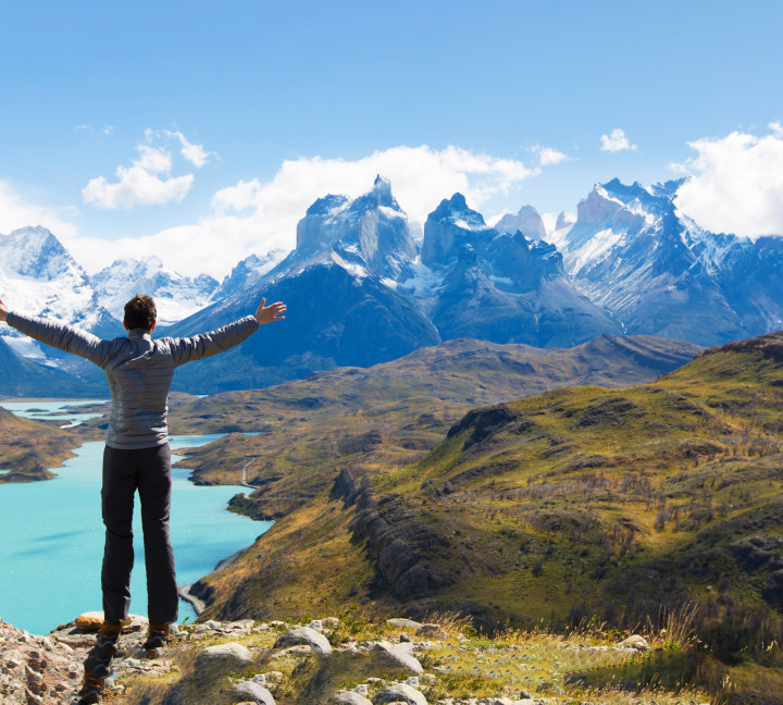 A guest stands on a cliff's edge with arms extended in front of clear light blue waters and lush green grass, with the  mountain peaks of Cuernos del Paine in the background. The photo was taken at Torres del Paine National Park, Chile.
