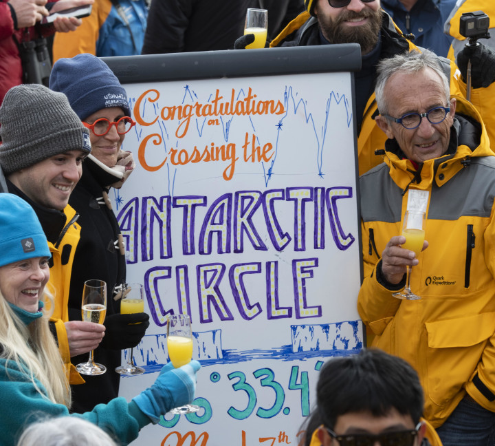 Guests celebrate crossing the Antarctic Circle with sparkling beverages and taking photos on the deck of the ship.