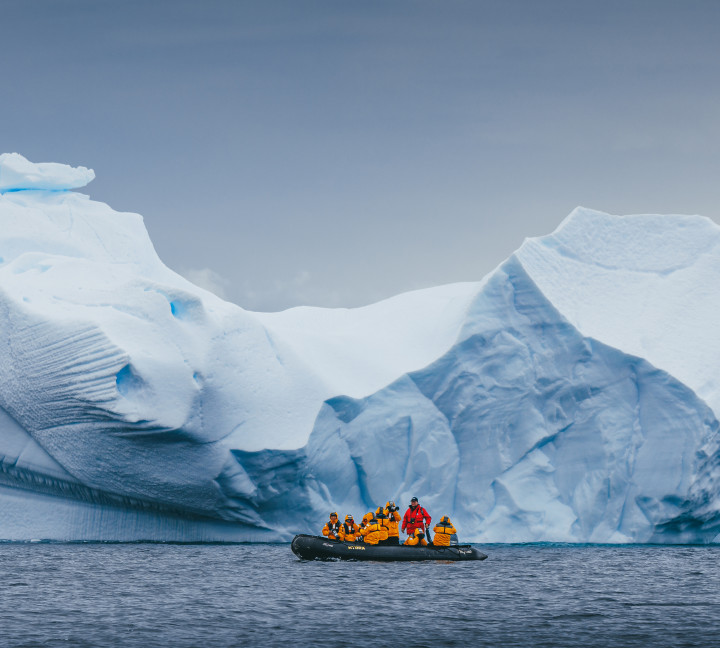 Zodiac boat in front of iceberg. Guests in yellow parkas can be seen taking photos while their Expedition Guide, in red, drives the boat.