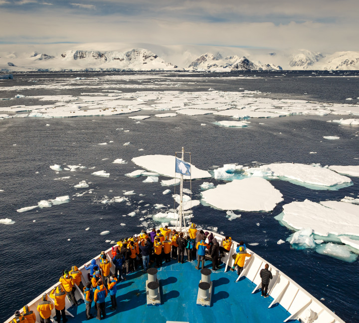 Guests in yellow parkas stand at the bow of their vessel as it cruises through sea ice in the Antarctic Peninsula.